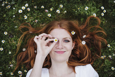 Smiling beautiful woman covering eye with daisy flower while lying on grass - EBBF02969