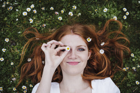 Lächelnde schöne Frau, die ihr Auge mit einer Gänseblümchenblüte bedeckt, während sie im Gras liegt - EBBF02969