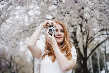 Young redhead woman photographing through camera at park - EBBF02971