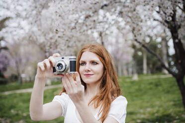 Beautiful redhead woman photographing through camera at park - EBBF02972
