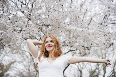 Glückliche junge Frau tanzt vor einem Mandelbaum und schaut weg - EBBF02975