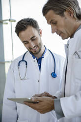 Männlicher Arzt im mittleren Erwachsenenalter, der mit einem Kollegen über ein digitales Tablet im Krankenhaus die Behandlung bespricht - BMOF00535