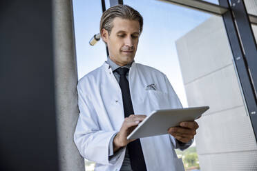 Männlicher Arzt im mittleren Erwachsenenalter, der ein digitales Tablet benutzt, während er sich auf eine Stange am Glasfenster eines Krankenhauses stützt - BMOF00536