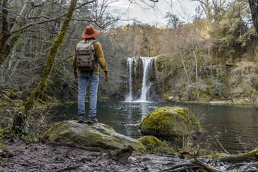 Mann schaut auf einen Wasserfall, während er auf einem Felsen am See steht - AFVF08467