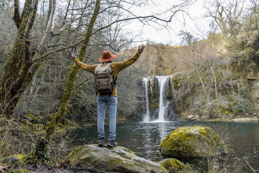 Mid adult man standing with arms outstretched in front of waterfall by lake - AFVF08468