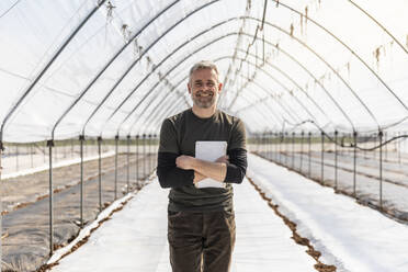 Lächelnder Bauer mit verschränkten Armen, der ein digitales Tablet in einem landwirtschaftlichen Feld hält - MCVF00755