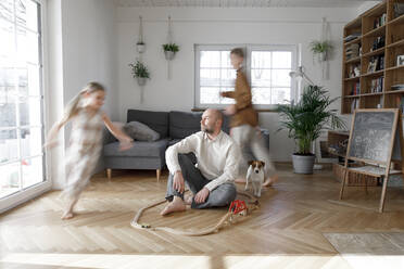 Siblings running around father sitting on floor in living room at home - KMKF01653
