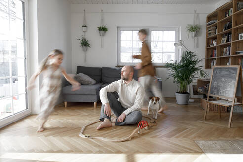 Geschwister rennen um den Vater herum, der zu Hause im Wohnzimmer auf dem Boden sitzt - KMKF01653