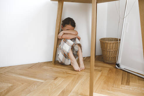 Deprimiertes kleines Mädchen sitzt unter dem Tisch auf dem Boden zu Hause - KMKF01657