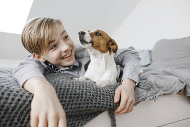 Smiling boy with arm around dog lying on bed at home - KMKF01659