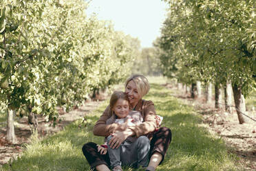 Happy mother with daughter sitting on grass in orchard - KMKF01668