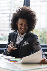 Glücklicher männlicher Jungunternehmer, der ein Dokument analysiert, während er einen Kaffee im Café trinkt - PNAF01275