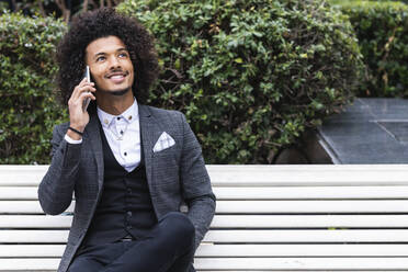 Männlicher Unternehmer, der auf einer Bank mit seinem Smartphone spricht und dabei wegschaut - PNAF01292