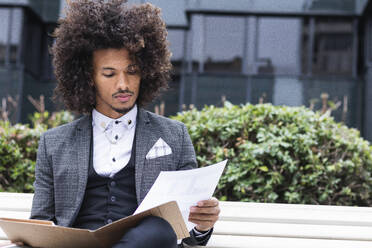 Junger männlicher Unternehmer, der auf einer Bank sitzend Dokumente in einer Akte prüft - PNAF01293