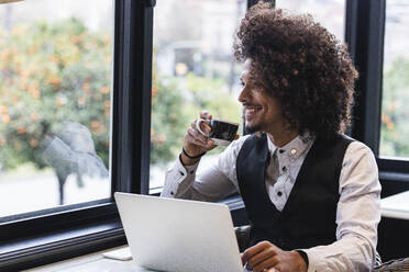 Smiling male entrepreneur with laptop having coffee while looking through window in cafe - PNAF01301