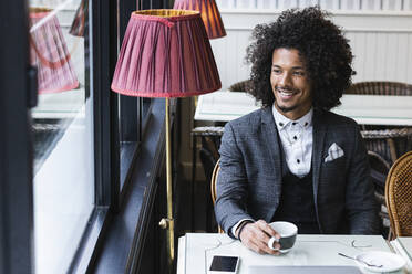 Lächelnder junger Unternehmer mit Kaffeetasse, der durch das Fenster eines Cafés schaut - PNAF01303