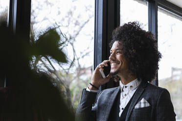 Smiling male entrepreneur looking through window while talking on mobile phone in cafe - PNAF01307