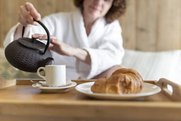 Frau gießt Kaffee in Tasse neben Croissant in Hotelzimmer - JPTF00730