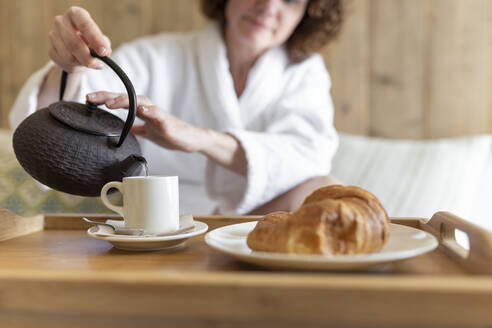 Frau gießt Kaffee in Tasse neben Croissant in Hotelzimmer - JPTF00730