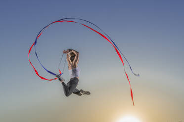 Female gymnast jumping with ribbons during sunset - STSF02896