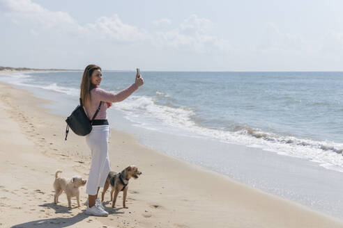 Frau mit Hunden spricht Selfie durch Smartphone am Strand während sonnigen Tag - JRVF00391