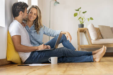 Happy couple with laptop looking at each other while sitting at home office - SBOF03670