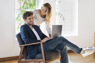 Smiling girlfriend looking at boyfriend using laptop while sitting on chair at home - SBOF03672