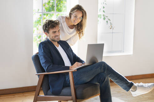 Mann benutzt Laptop, während er bei seiner Freundin zu Hause auf dem Stuhl sitzt - SBOF03673