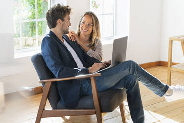 Man sitting on chair with laptop looking at girlfriend at home - SBOF03675