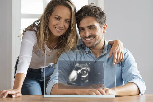 Glückliches Paar betrachtet Ultraschallbild auf transparentem Bildschirm zu Hause - SBOF03694