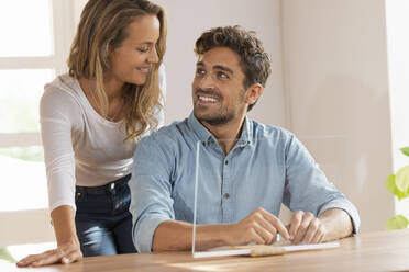 Happy couple looking at each other sitting with transparent screen at home - SBOF03695