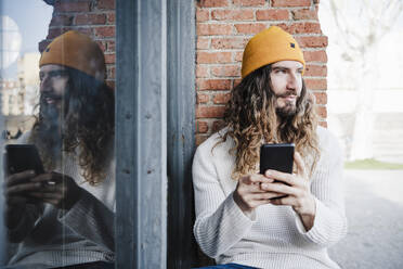 Bearded man with mobile phone looking away while sitting in front of old wall - EBBF02996