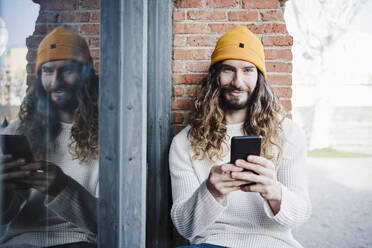 Lächelnder Mann mit Mobiltelefon vor einer Wand sitzend - EBBF02997