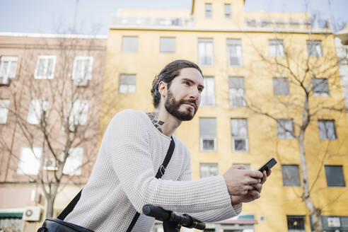 Hipster-Mann mit Smartphone, der sich auf einen Elektroroller stützt und wegschaut - EBBF03001