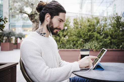 Männlicher Hipster mit Einwegbecher, der während eines Videogesprächs in einem Straßencafé ein digitales Tablet berührt - EBBF03012