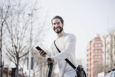 Glücklicher männlicher Hipster, der sein Smartphone auf einem Motorroller hält - EBBF03025