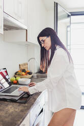 Mature woman in shirt working on laptop in kitchen at home - MRRF00964