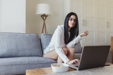 Frau benutzt einen Laptop, während sie auf dem Sofa im Wohnzimmer sitzt - MRRF00966