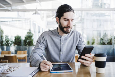Geschäftsmann mit Grafiktablett und wiederverwendbarem Kaffeebecher bei der Nutzung eines Smartphones im Büro - EBBF03028