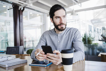 Contemplative male entrepreneur looking away while sitting with wireless technologies at work place - EBBF03029