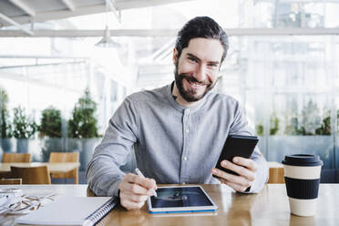 Lächelnder Geschäftsmann mit digitalem Tablet und Smartphone am Arbeitsplatz - EBBF03030