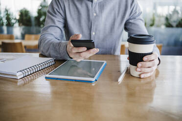 Mittlerer erwachsener Geschäftsmann mit wiederverwendbarem Kaffeebecher und Smartphone auf dem Schreibtisch im Büro - EBBF03036