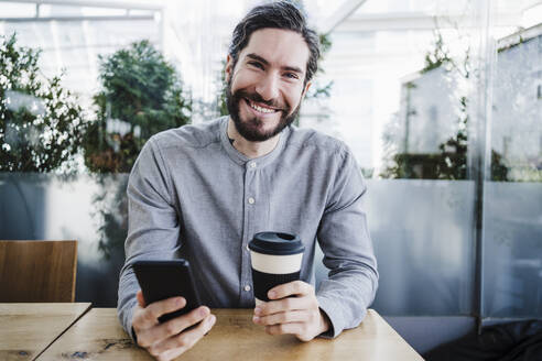 Glücklicher männlicher Geschäftsmann mit Smartphone und wiederverwendbarem Kaffeebecher im Büro - EBBF03044