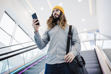 Mann mit langen Haaren, der ein Mobiltelefon hält, während er sich auf einer Rolltreppe bewegt - EBBF03058