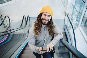 Lächelnder Mann mit Smartphone, der auf einer Rolltreppe nach oben fährt - EBBF03059