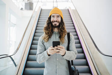 Glücklicher Mann mit Laptoptasche und Smartphone vor einer Rolltreppe - EBBF03062