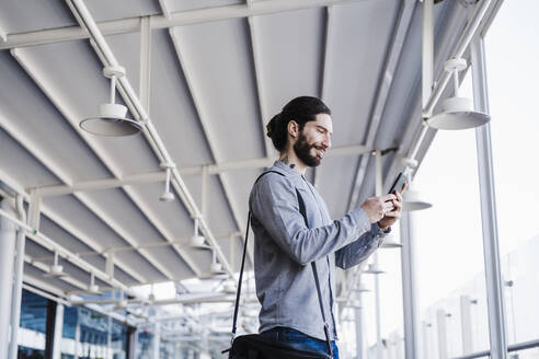 Smiling businessman with laptop bag using mobile phone in office balcony - EBBF03075