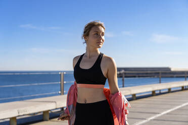 Beautiful female jogger removing jacket while looking away during sunny day - AMPF00186