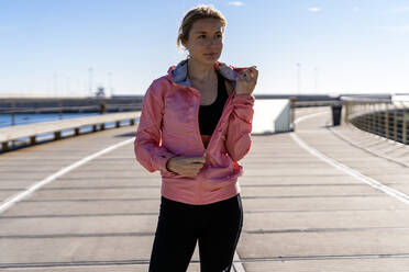 Female athlete unzipping jacket while standing on boardwalk - AMPF00187