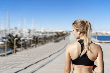 Weiblicher Sportler mit In-Ear-Kopfhörern, der wegschaut, während er an einem sonnigen Tag auf der Strandpromenade steht - AMPF00191